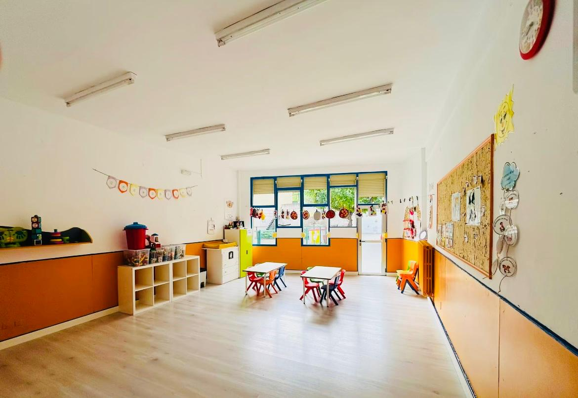 Aula luminosa con zócalo naranja, mesas y sillas infantiles y corcho decorado