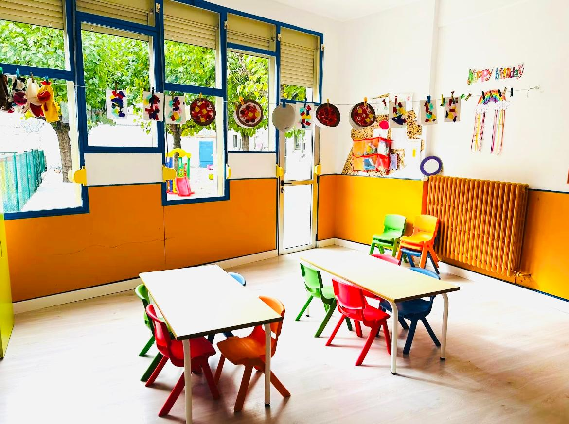 Aula con ventanales, manualidades colgadas y mesas con sillas de colores