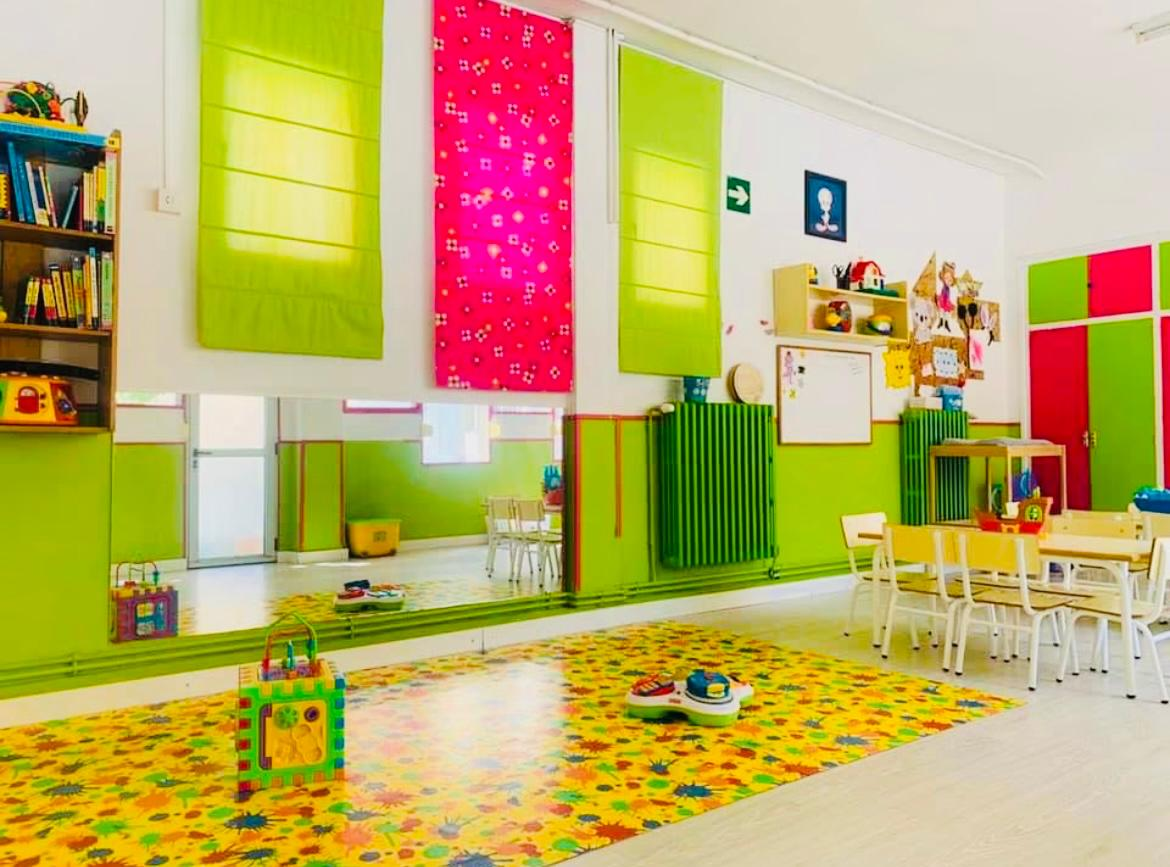 Aula amplia con cortinas verdes y rosa, alfombra colorida y mesas infantiles