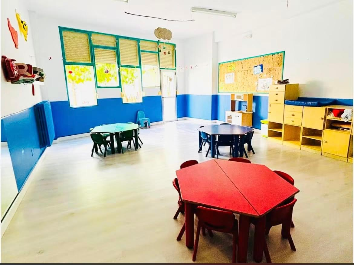 Aula con mesas hexagonales rojas y verdes, sillas infantiles y cocinita de juguete